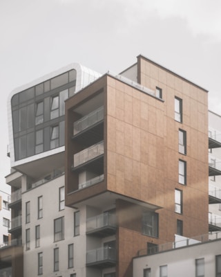 A modern multi-story residential building with a combination of materials, including glass, metal, and brown paneling. The architecture features a mix of rectangular shapes with several protruding balconies, providing contrast between smooth white and textured brown surfaces. Large windows dominate the upper floors, allowing for natural light to enter.
