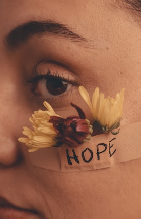 A close-up of a person's face with a focus on the eye, showing a band-aid with the word 'HOPE' written on it. Attached to the band-aid are two yellow flowers and a red flower bud.