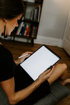 A person sitting on a chair holding a large tablet with a blank screen, using a stylus pen. In the background, there is a bookshelf partially filled with books.