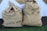 Two burlap sacks filled with coffee, labeled with text indicating they are products of El Salvador from the 2017/2018 harvest. The sacks have printing in black ink, including the words 'QUETA SHG' and crop details. They are sitting on a surface with a white background cloth and a green carpet.