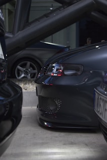 A close-up view of three cars parked closely together in a dimly lit area. The front bumper of one car is prominently visible, featuring a mesh detail and sleek, dark design. In the background, another car with a similar dark finish is partially visible, highlighting its wheel and body structure. The setting seems to be an industrial or urban environment with metal beams and a concrete surface.