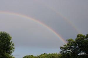 A double rainbow arches across a cloudy sky, with the vibrant colors contrasting against the backdrop. Below, lush green trees create a natural frame, adding depth and tranquility to the scene. The soft light suggests the aftermath of a rain shower, enhancing the atmospheric quality of the landscape.