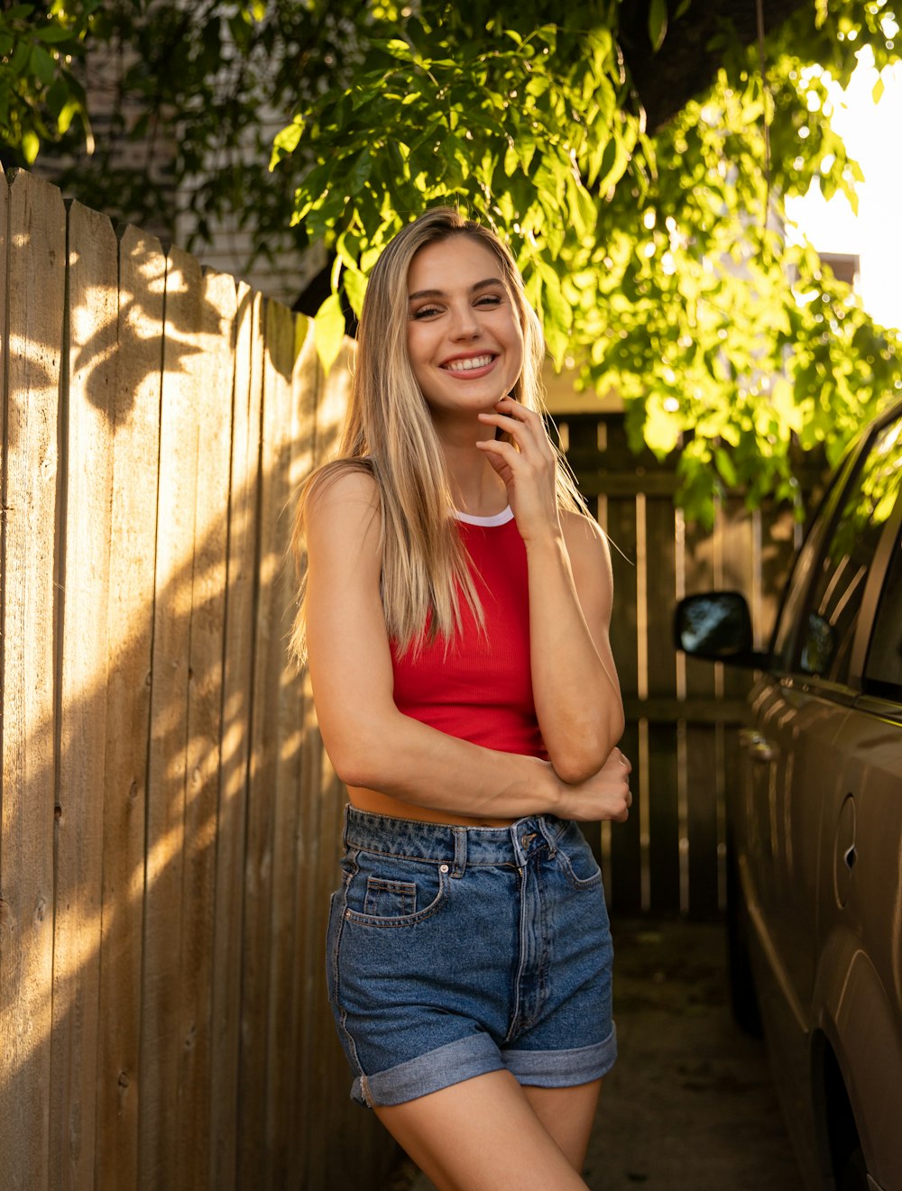 woman in red tank top and blue denim jeans standing beside brown wooden fence