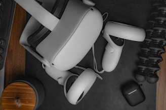 Various technology-related items are placed on a dark surface. A virtual reality headset with controllers is centrally located. Nearby, there is a black case, a tripod, and partially visible accessories including a wooden coaster or speaker.