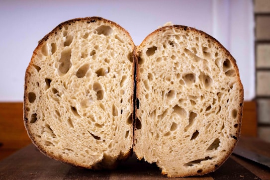 A cross-section of a loaf of bread, displaying an open and airy crumb structure with a well-baked crust. The bread is placed on a wooden surface against a soft, neutral background.