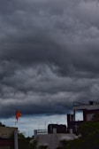 Dark, heavy clouds fill the sky above a residential area. Buildings with flat roofs are visible in the foreground, including a building with a bright orange flag flying from a pole. The atmosphere suggests approaching rain or a storm.