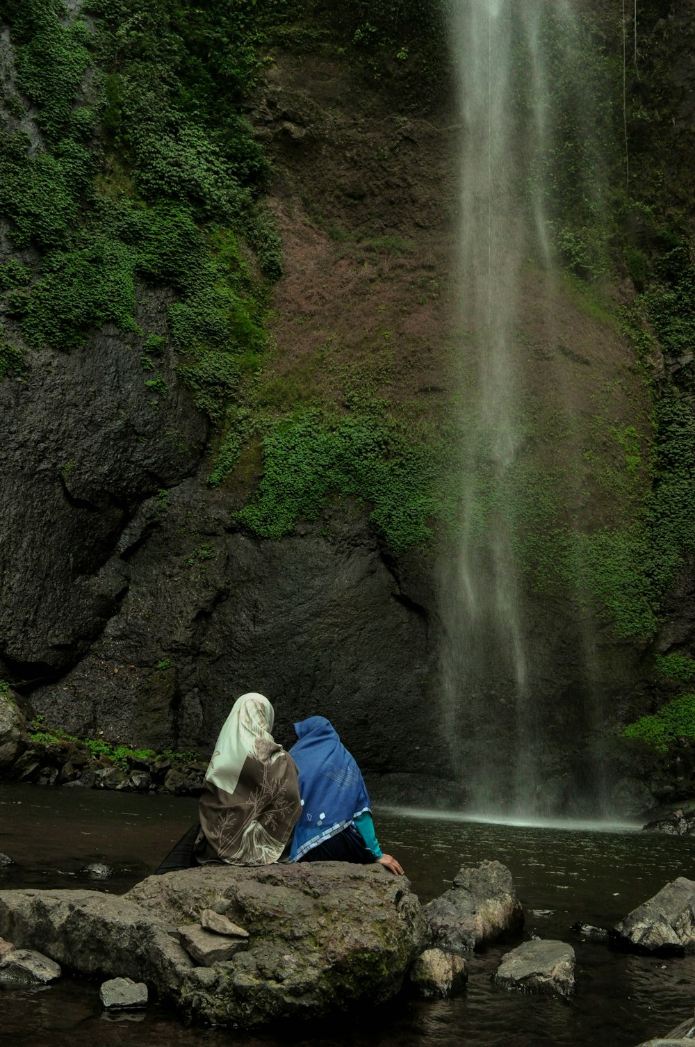 woman in blue jacket sitting on rock near waterfalls during daytime