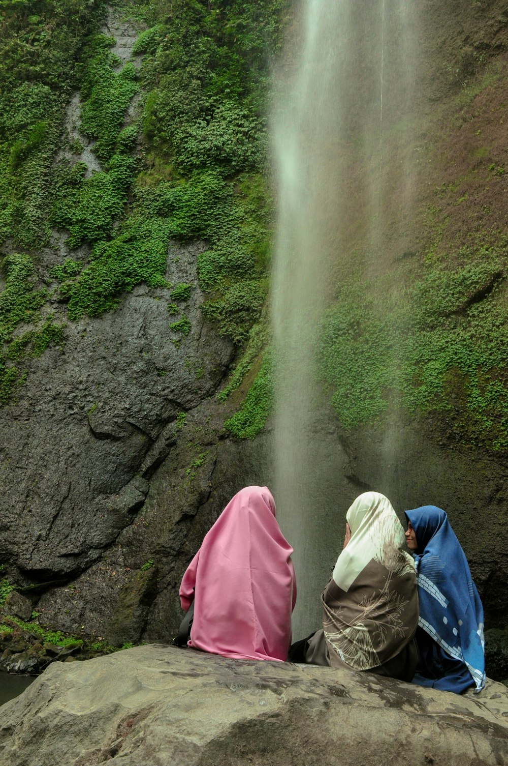 person in white and blue hoodie sitting on rock near waterfalls during daytime