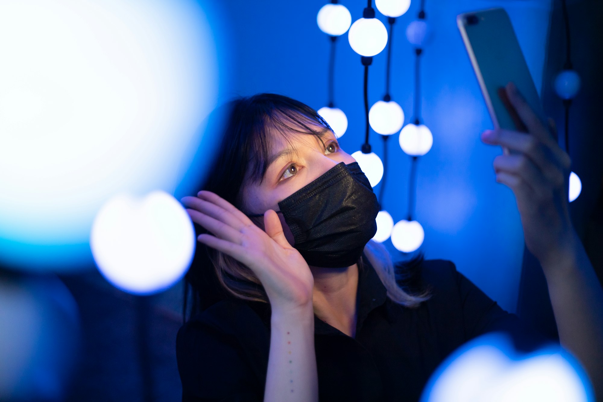 A woman wearing a mask uses her smart phone to video call at night surrounded by glowing blue lights.