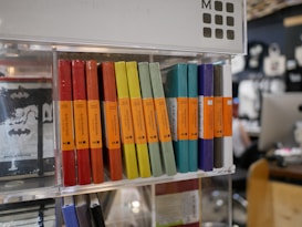A display shelf filled with various colored Moleskine notebooks, neatly arranged side by side. Each notebook has a vibrant colored cover including shades of red, yellow, green, turquoise, blue, and gray. Orange labels are attached to the spines, indicating that these are ruled notebooks. The background features a blurred scene of a store environment with shelves and other customers.
