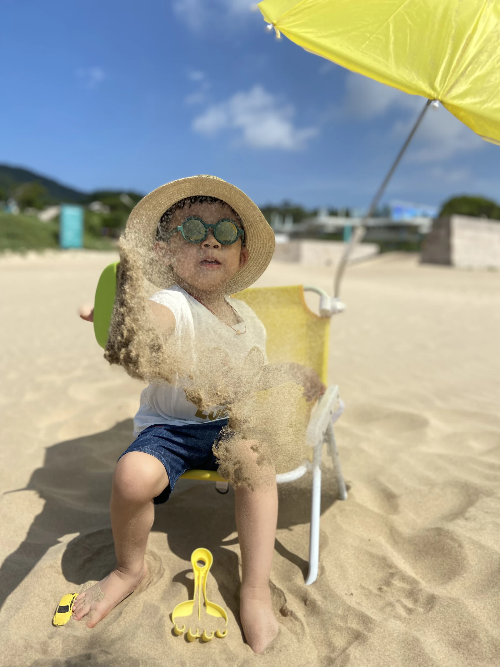 A young child in a sun hat and sunglasses sitting in the shade of a beach umbrella on a bright summer day