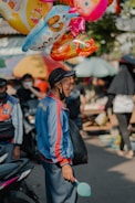 A street vendor in a colorful jacket stands holding a bunch of vibrant, cartoon-themed balloons. He appears to be in a busy outdoor market with people and motorbikes in the background. The vendor carries a black bag and wears a cap.