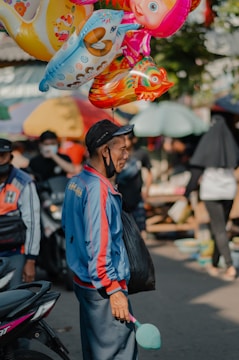 A street vendor in a colorful jacket stands holding a bunch of vibrant, cartoon-themed balloons. He appears to be in a busy outdoor market with people and motorbikes in the background. The vendor carries a black bag and wears a cap.