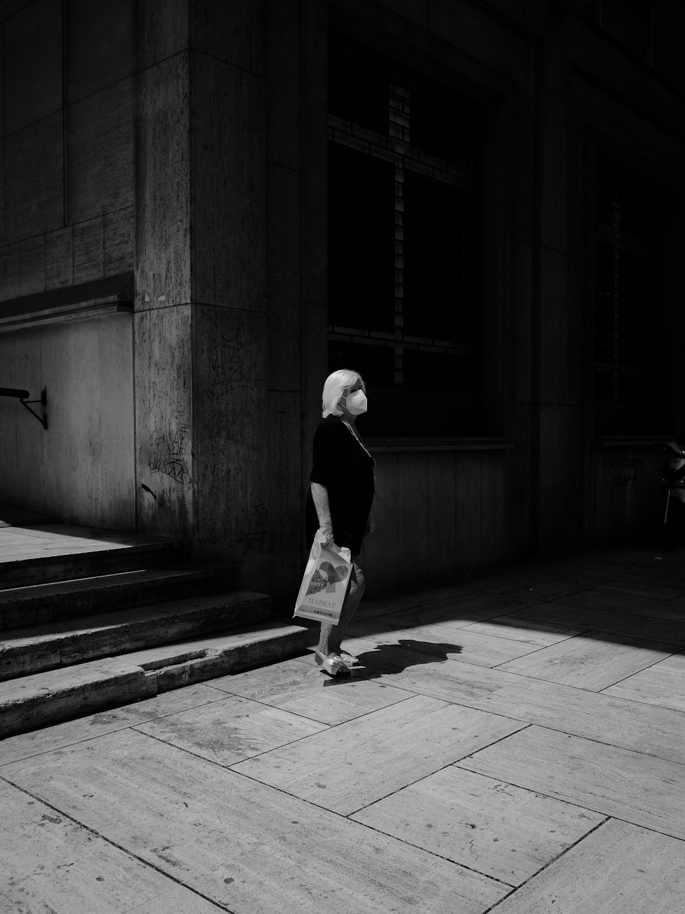Foto in scala di grigi di uomo in camicia nera e pantaloni che camminano sul marciapiede