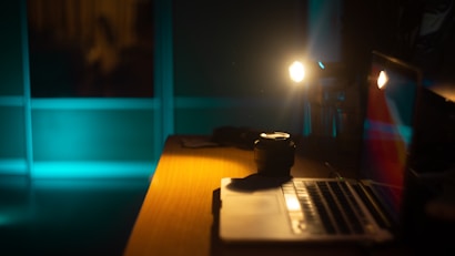 A dimly lit workspace features a laptop on a wooden desk, with its screen partially visible and reflecting ambient colors. A camera lens rests next to the laptop, while two muted lamps cast soft light, creating a warm and intimate atmosphere. The background is a window with a faint blue hue, suggesting nighttime.