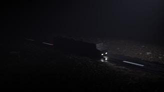 A dark, nighttime scene featuring a vehicle with headlights on, traveling on a road. The surroundings are dimly lit, creating an eerie and mysterious atmosphere. The road surface appears wet and reflective.