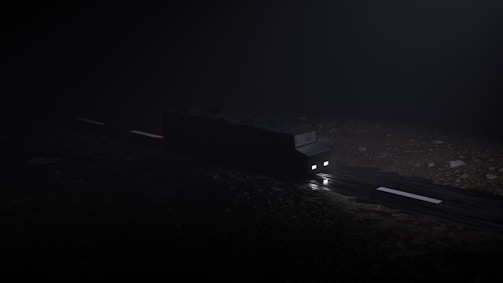 A dark, nighttime scene featuring a vehicle with headlights on, traveling on a road. The surroundings are dimly lit, creating an eerie and mysterious atmosphere. The road surface appears wet and reflective.