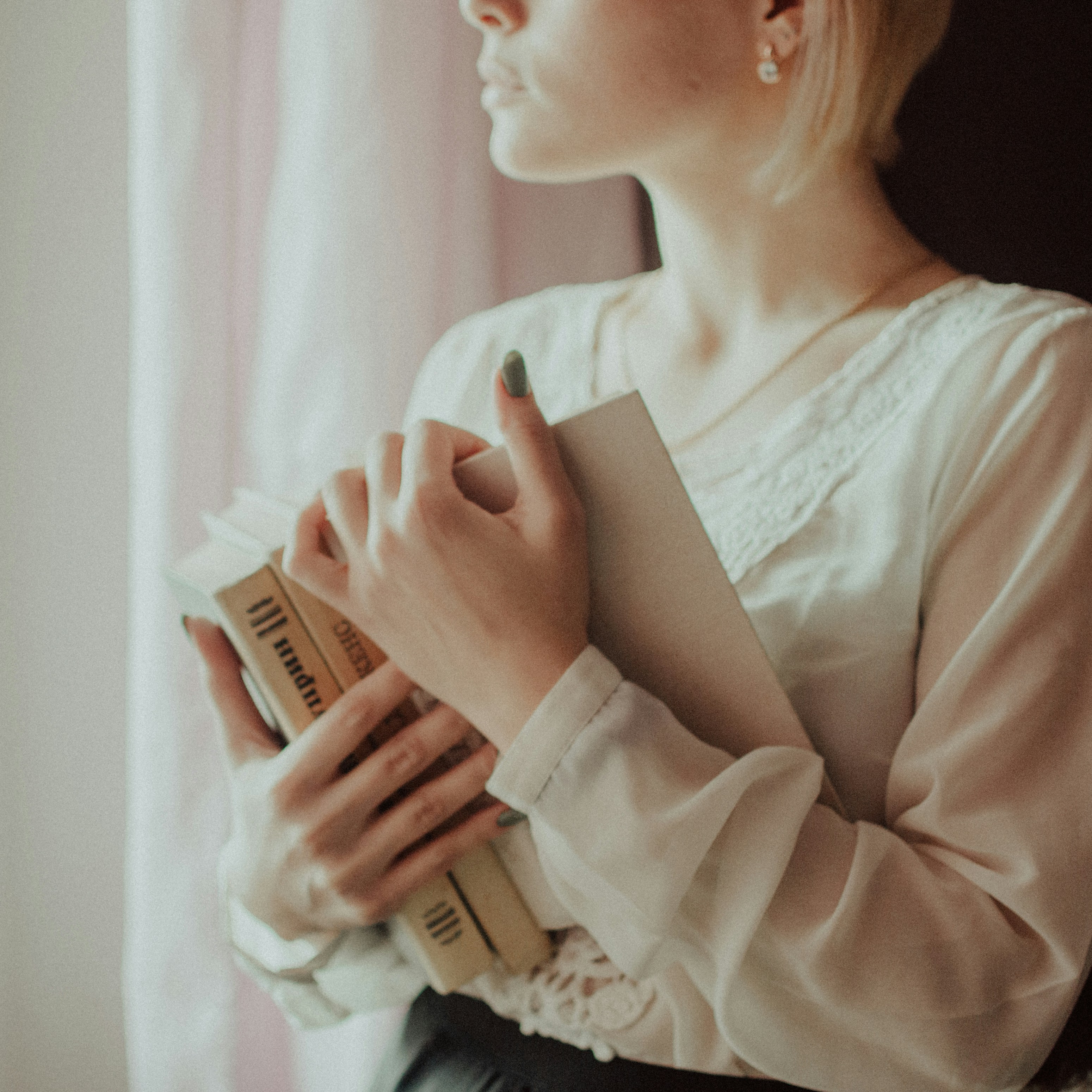 A woman stands by a window, holding a book, with natural light illuminating her thoughtful expression.