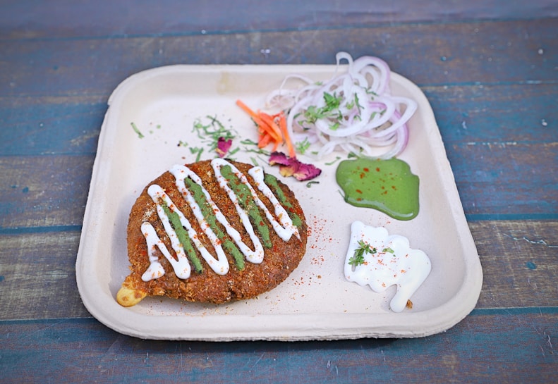 A fried patty topped with white and green sauces is placed on a compostable plate. Accompanied by thinly sliced onions, carrots, and sprigs of herbs, it is garnished with small portions of green chutney and white yogurt dip. The background features a rustic, blue-toned wooden surface, adding a vibrant contrast to the presentation.