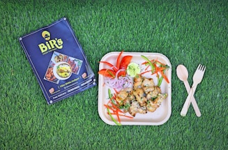 A square plate with grilled chicken pieces, garnished with fresh herbs and accompanied by sliced vegetables including tomatoes, red onions, carrots, and green chili peppers, arranged neatly on a green grass background. To the left of the plate is a restaurant menu, and on the right are wooden fork and spoon placed crossed over each other.