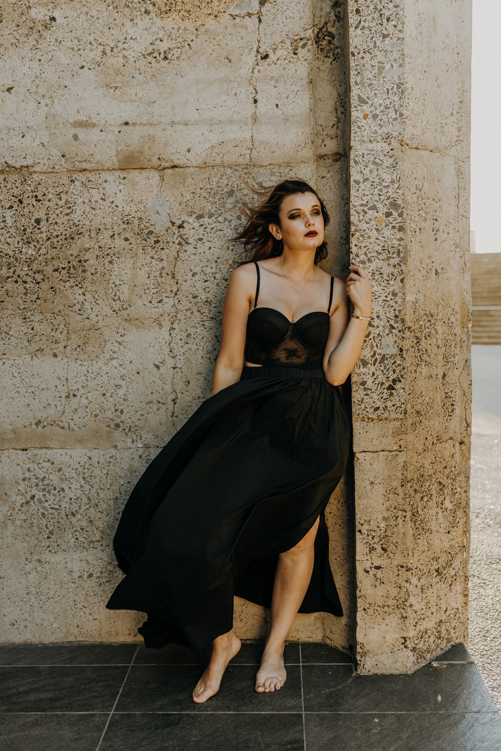 woman in black spaghetti strap dress leaning on gray concrete wall