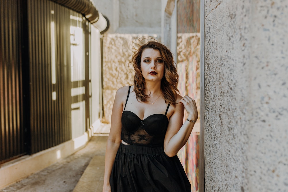 woman in black spaghetti strap dress leaning on white concrete wall during daytime