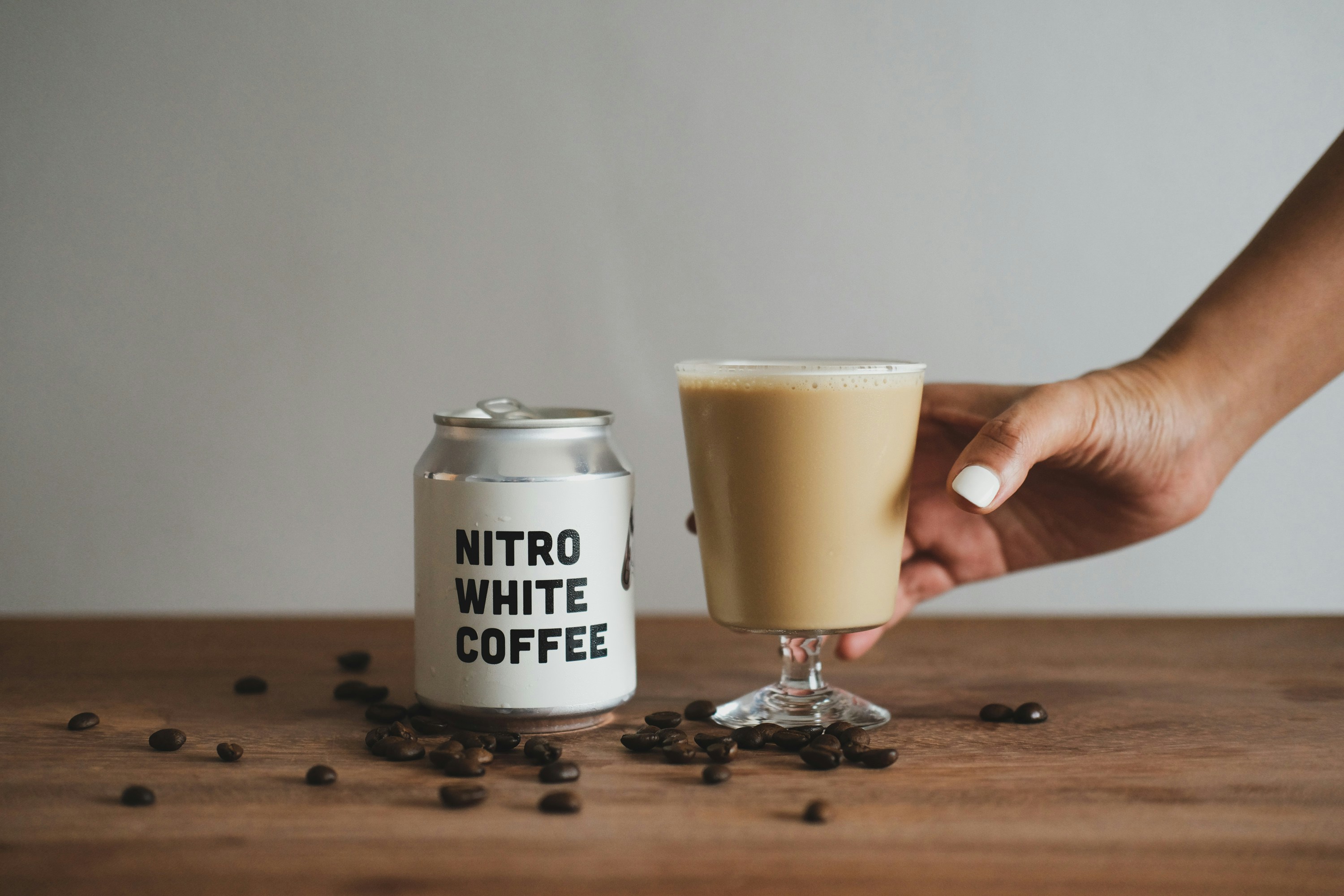A can of Pushers nitro flat white / cafe latte sitting on a wooden table with coffee beans strewn about and a hand picking up the glass.
