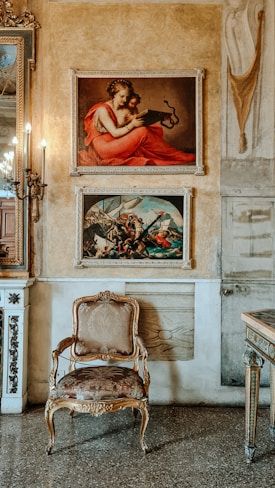 An elegant room with ornate decor, featuring two framed paintings on a textured wall. The upper painting depicts a figure in a red garment with an open book, while the lower painting presents a dynamic scene with multiple figures. A classic baroque-style armchair with intricate patterns and a polished wooden frame is placed below the paintings. The mirror to the left and part of the console table to the right enhance the refined ambiance, complemented by soft lighting from wall-mounted candleholders.