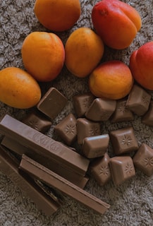 A collection of ripe apricots with a smooth, vibrant orange and red skin resting on a textured surface. Below them, a variety of chocolate pieces, including rectangular chocolate bars and small square chocolates with embossed star patterns, are arranged in a casual cluster.