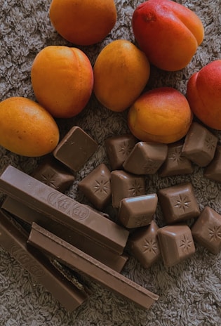 A collection of ripe apricots with a smooth, vibrant orange and red skin resting on a textured surface. Below them, a variety of chocolate pieces, including rectangular chocolate bars and small square chocolates with embossed star patterns, are arranged in a casual cluster.