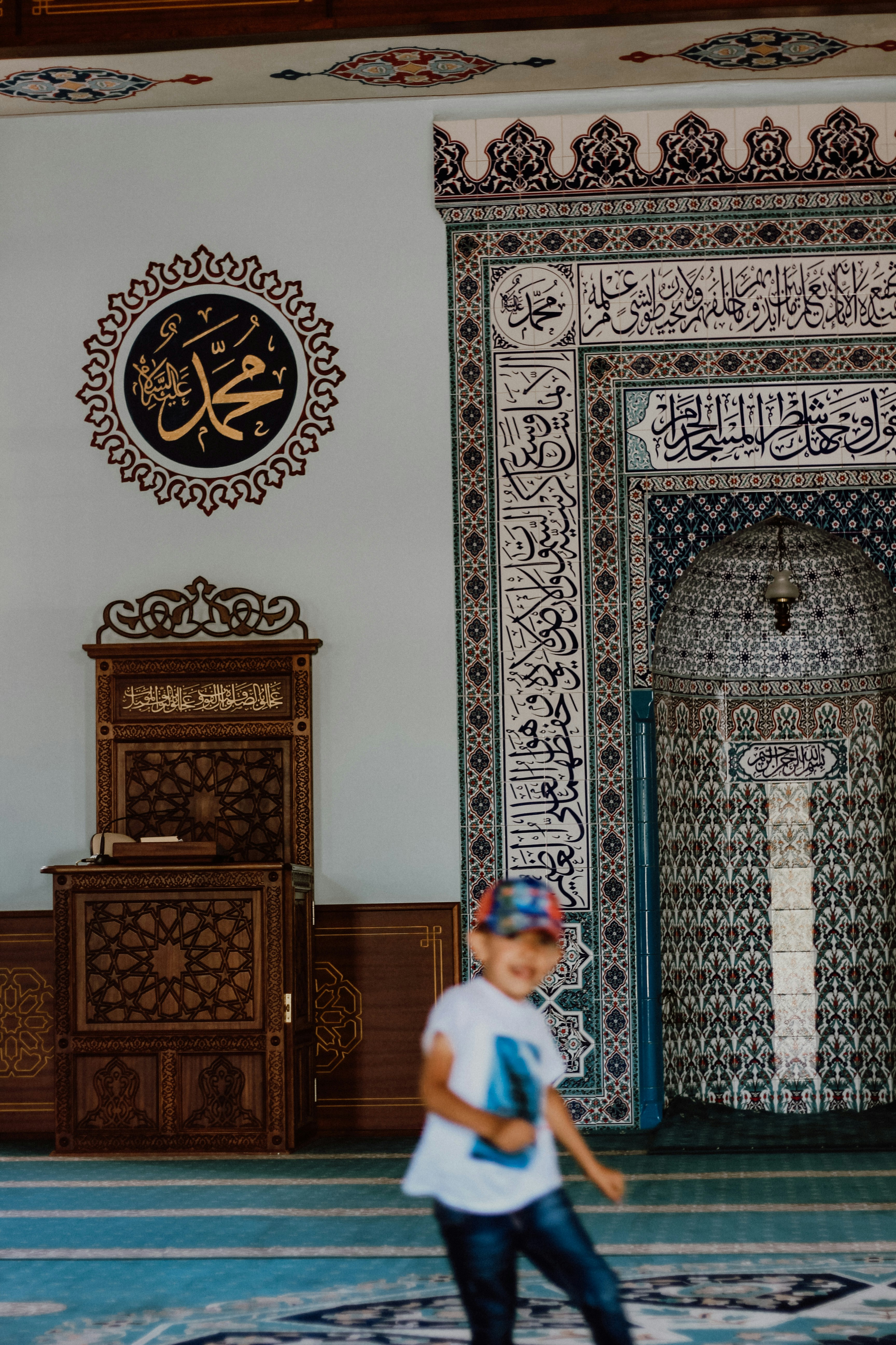 A mosque interior features intricate calligraphy and geometric patterns on the walls. A wooden lectern with ornate carvings sits to the left. A child wearing a white shirt and a colorful cap is in motion in the foreground.