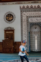 A mosque interior features intricate calligraphy and geometric patterns on the walls. A wooden lectern with ornate carvings sits to the left. A child wearing a white shirt and a colorful cap is in motion in the foreground.