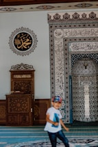A mosque interior features intricate calligraphy and geometric patterns on the walls. A wooden lectern with ornate carvings sits to the left. A child wearing a white shirt and a colorful cap is in motion in the foreground.