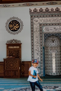 A mosque interior features intricate calligraphy and geometric patterns on the walls. A wooden lectern with ornate carvings sits to the left. A child wearing a white shirt and a colorful cap is in motion in the foreground.
