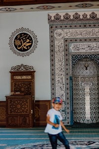 A mosque interior features intricate calligraphy and geometric patterns on the walls. A wooden lectern with ornate carvings sits to the left. A child wearing a white shirt and a colorful cap is in motion in the foreground.