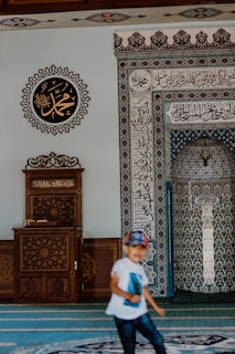 A mosque interior features intricate calligraphy and geometric patterns on the walls. A wooden lectern with ornate carvings sits to the left. A child wearing a white shirt and a colorful cap is in motion in the foreground.