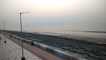 A serene beach scene during sunset with a calm sea and a soft, hazy sky. The sun appears as a small glowing dot near the horizon. A wide promenade with patterned tiles runs parallel to the beach with a few scattered individuals walking. Streetlights line the promenade, and the beach itself is sparsely populated.