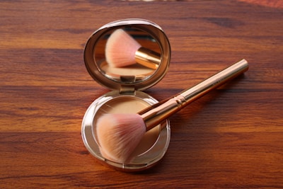 A makeup brush with a pink, fluffy bristle head rests inside an open circular compact mirror case on a wooden surface. The compact case has a reflective interior and contains a beige powder.