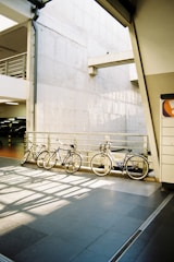 Three bicycles are lined up against a metal railing in a modern urban parking area. The setting includes a concrete wall with geometric architectural elements and diffused natural light, which creates interesting shadow patterns on the shiny tiled floor.