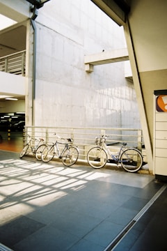 Three bicycles are lined up against a metal railing in a modern urban parking area. The setting includes a concrete wall with geometric architectural elements and diffused natural light, which creates interesting shadow patterns on the shiny tiled floor.