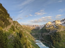 A picturesque mountain landscape featuring snow-capped peaks and lush green valleys. A winding road leads through the valley, beside a small reflective lake. The sky is clear with scattered clouds, enhancing the tranquil and serene atmosphere.