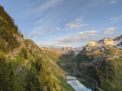A picturesque mountain landscape featuring snow-capped peaks and lush green valleys. A winding road leads through the valley, beside a small reflective lake. The sky is clear with scattered clouds, enhancing the tranquil and serene atmosphere.