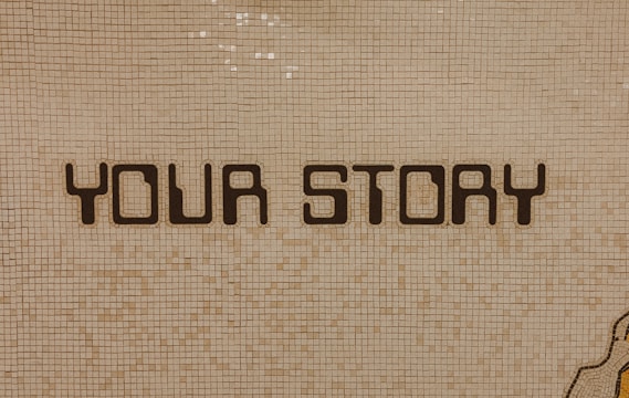 The image contains a mosaic wall with the words 'YOUR STORY' written in bold, dark letters. The tiles are small and mostly uniform, giving a textured appearance to the background.