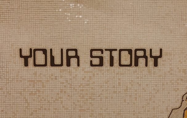 The image contains a mosaic wall with the words 'YOUR STORY' written in bold, dark letters. The tiles are small and mostly uniform, giving a textured appearance to the background.