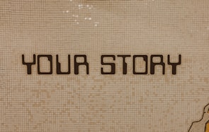 The image contains a mosaic wall with the words 'YOUR STORY' written in bold, dark letters. The tiles are small and mostly uniform, giving a textured appearance to the background.