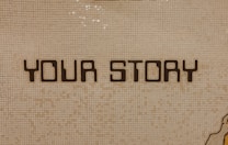 The image contains a mosaic wall with the words 'YOUR STORY' written in bold, dark letters. The tiles are small and mostly uniform, giving a textured appearance to the background.