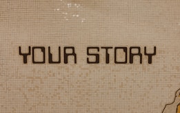The image contains a mosaic wall with the words 'YOUR STORY' written in bold, dark letters. The tiles are small and mostly uniform, giving a textured appearance to the background.