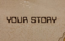 The image contains a mosaic wall with the words 'YOUR STORY' written in bold, dark letters. The tiles are small and mostly uniform, giving a textured appearance to the background.