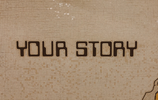 The image contains a mosaic wall with the words 'YOUR STORY' written in bold, dark letters. The tiles are small and mostly uniform, giving a textured appearance to the background.