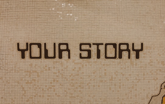 The image contains a mosaic wall with the words 'YOUR STORY' written in bold, dark letters. The tiles are small and mostly uniform, giving a textured appearance to the background.